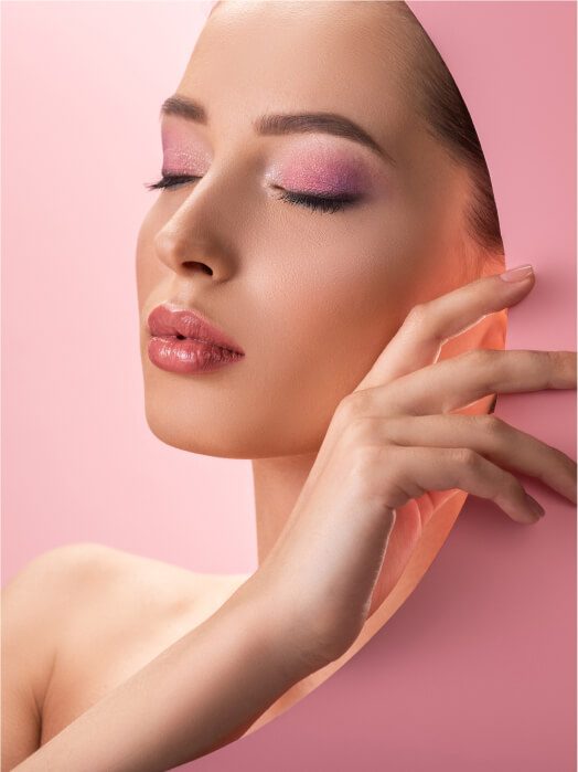 Femme en profil avec maquillage rose et peau lisse, main touchant délicatement le visage sur fond rose pastel