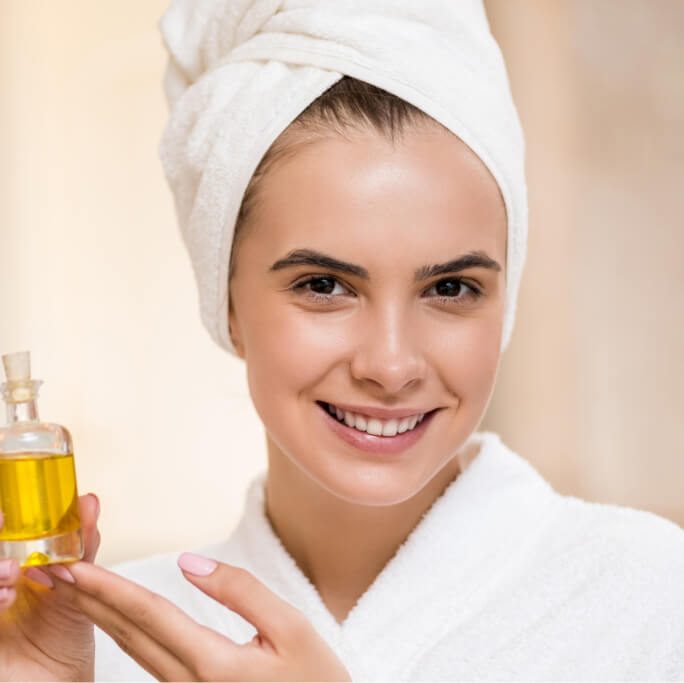 Femme souriante enveloppée dans une serviette blanche tenant une bouteille d'huile de soin