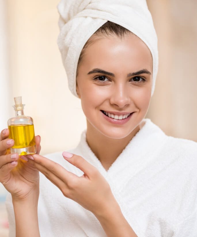 Jeune femme souriante enveloppée dans une serviette blanche tenant une bouteille d'huile dorée pour soins esthétiques