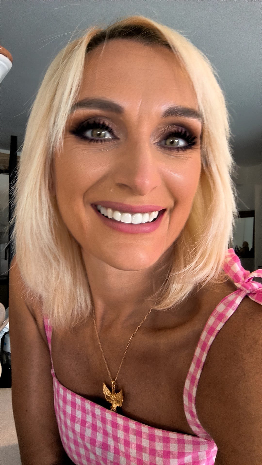 Femme blonde souriante portant un haut rose à carreaux avec un pendentif doré en forme d'oiseau